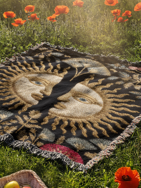 Jacquard-woven cotton blanket featuring twin celestial suns—two serene faces meeting at center—with radiant golden rays on a charcoal ground, bordered by folkloric botanicals and fan blossoms in sage, sky, and garnet. Crafted from 100% soft cotton yarn; the artwork is woven, not printed. Reversible with a color-negative back and finished with a protective 2-inch multicolor fringe. Sizes: 52×37, 50×60, 60×80 for wall, sofa, or queen-bed layering.
